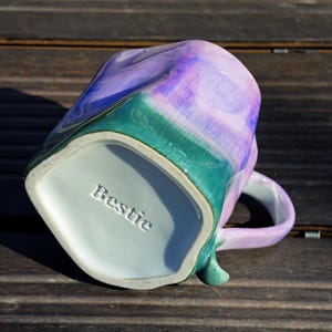 May include: A ceramic mug with a unique, irregular shape. The mug features a blend of colors, including purple, blue, and green, with a glossy finish. The word "Bestie" is embossed on the bottom of the mug. The mug has a curved handle.