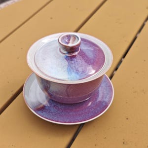 May include: A ceramic tea set with a purple and white glaze. The set includes a lidded bowl and a matching saucer. The lid has a small, rounded handle. The colors blend together, creating a unique and artistic appearance.