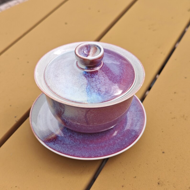 May include: A ceramic tea set with a purple and white glaze. The set includes a lidded bowl and a matching saucer. The lid has a small, rounded handle. The colors blend together, creating a unique and artistic appearance.