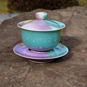 May include: A ceramic tea set with a lid, cup, and saucer. The set features a gradient of colors, including teal, purple, and white. The cup and saucer are round, and the lid has a small handle. The set is placed on a stone surface.