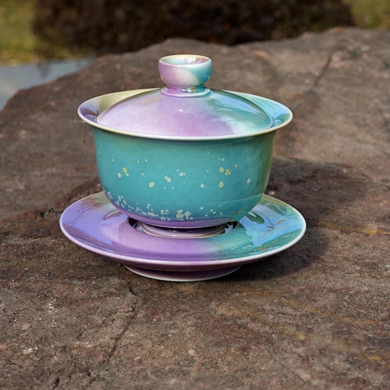 May include: A ceramic tea set with a lid, cup, and saucer. The set features a gradient of colors, including teal, purple, and white. The cup and saucer are round, and the lid has a small handle. The set is placed on a stone surface.