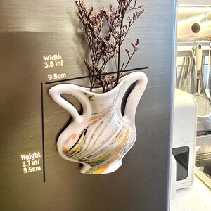 Op de afbeelding: Witte keramische vaas met een uniek, gebogen ontwerp, 9,5 cm breed en 9,5 cm hoog, gevuld met gedroogde bloemen. De vaas heeft kleurrijke verfstrepen en is bevestigd aan een metalen oppervlak.
