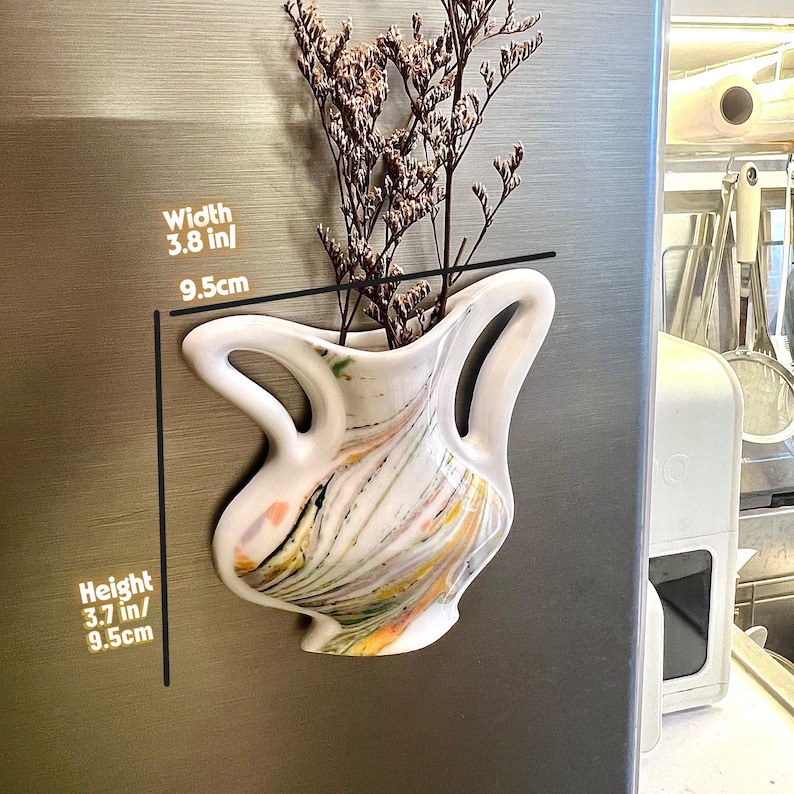 Op de afbeelding: Witte keramische vaas met een uniek, gebogen ontwerp, 9,5 cm breed en 9,5 cm hoog, gevuld met gedroogde bloemen. De vaas heeft kleurrijke verfstrepen en is bevestigd aan een metalen oppervlak.