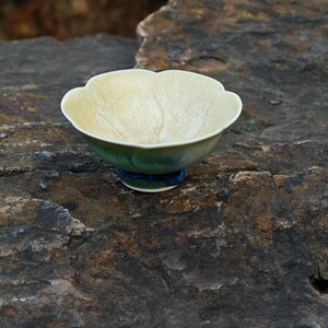 May include: A ceramic tea bowl with a unique, scalloped rim. The interior is a pale yellow with subtle veining, while the exterior transitions from green to blue. The bowl is approximately 3 inches in diameter.