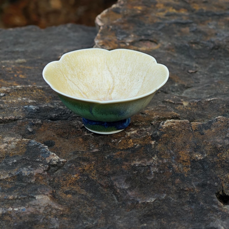 May include: A ceramic tea bowl with a unique, scalloped rim. The interior is a pale yellow with subtle veining, while the exterior transitions from green to blue. The bowl is approximately 3 inches in diameter.
