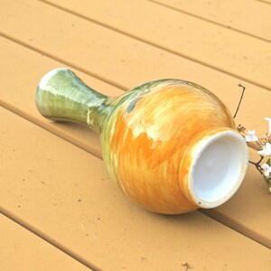K&ouml;nnte beinhalten: Eine Keramikvase mit bauchigem K&ouml;rper und schmalem Hals. Die Vase ist mit einer Mischung aus orange, gelb und gr&uuml;nen Farbt&ouml;nen bemalt. Die &Ouml;ffnung ist wei&szlig;. Die Vase liegt auf einer Holzoberfl&auml;che.