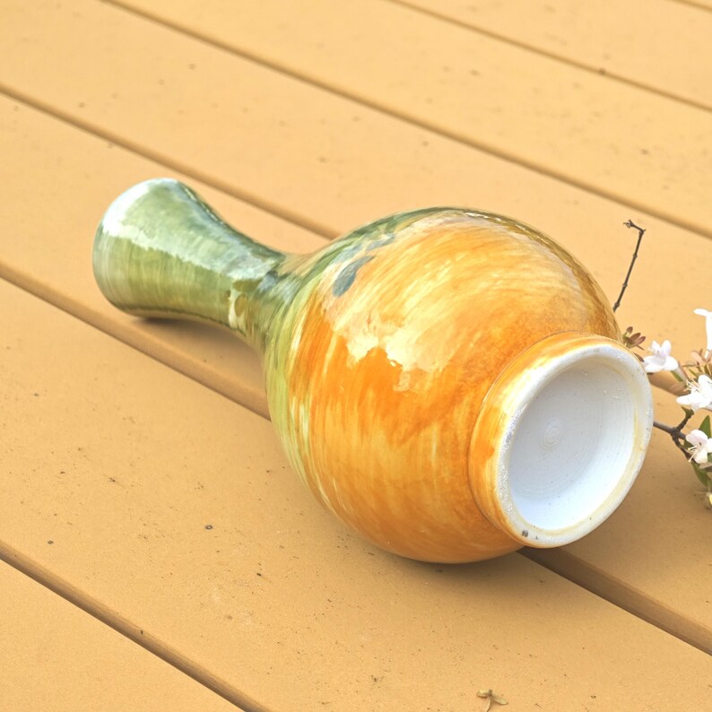 K&ouml;nnte beinhalten: Eine Keramikvase mit bauchigem K&ouml;rper und schmalem Hals. Die Vase ist mit einer Mischung aus orange, gelb und gr&uuml;nen Farbt&ouml;nen bemalt. Die &Ouml;ffnung ist wei&szlig;. Die Vase liegt auf einer Holzoberfl&auml;che.