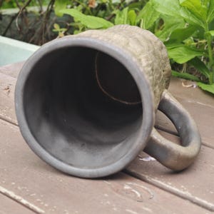 Puede incluir: Una taza de cer&aacute;mica con un interior gris carb&oacute;n oscuro y una superficie exterior texturizada de color marr&oacute;n claro. La taza cuenta con un asa gruesa y curva y un borde circular ancho, mostrando el contraste entre la pared interior lisa y oscura.