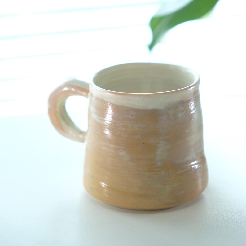 May include: A handmade ceramic mug with a light brown and cream color scheme. The mug has a rounded body, a sturdy handle, and a smooth, glossy finish. The mug is suitable for coffee, tea, or other beverages.
