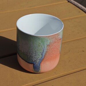 Pu&ograve; includere: Una tazza cilindrica in ceramica con interno bianco ed esterno colorato. L'esterno presenta una miscela di colori, tra cui corallo, verde e blu, creando un design artistico unico. La tazza viene probabilmente utilizzata per bere bevande.