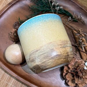 May include: A ceramic tumbler with a gradient design, transitioning from blue at the rim to yellow and brown at the base. The handmade mug is set on a wooden tray with decorative elements, including pine cones and dried foliage.