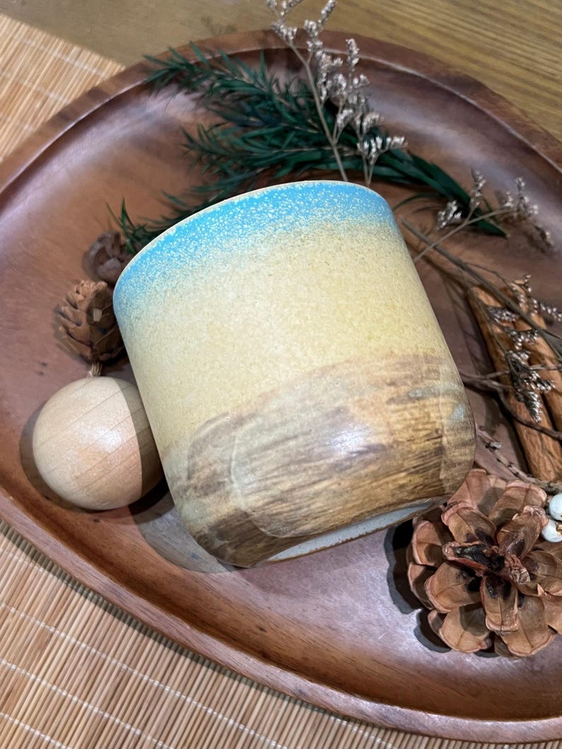 May include: A ceramic tumbler with a gradient design, transitioning from blue at the rim to yellow and brown at the base. The handmade mug is set on a wooden tray with decorative elements, including pine cones and dried foliage.