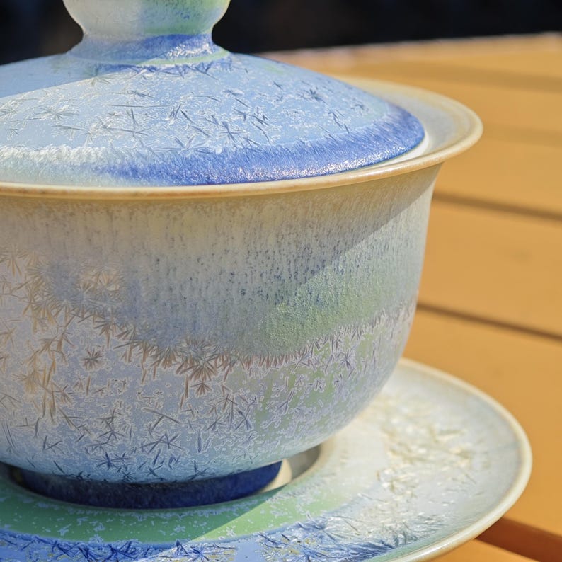 May include: A ceramic tea set with a light blue and white speckled glaze. The set includes a lidded bowl and a matching saucer. The lid and the rim of the bowl have a darker blue band. The set is on a wooden surface.