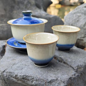 May include: A ceramic tea set with a blue and cream color scheme. The set includes a lidded gaiwan, a saucer, and two teacups. The teacups and gaiwan have a blue base and rim, with a cream-colored body. The set is displayed on a gray rock.