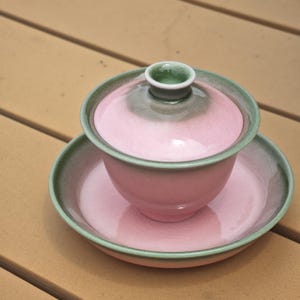 Puede incluir: Un juego de t&eacute; gaiwan de cer&aacute;mica compuesto por un cuenco, una tapa y un platillo. Las piezas presentan un centro rosa suave con un acabado de esmalte craquelado, que transiciona hacia un borde verde salvia apagado. La tapa tiene un pomo central.