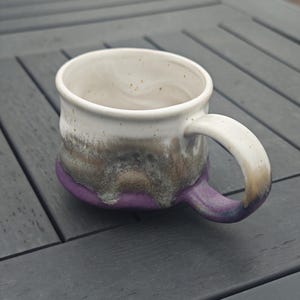 Op de afbeelding: Een handgemaakte keramische mok met een witte binnenkant en een oor. De buitenkant heeft een kleurengradi&euml;nt, van wit naar bruin, grijs en paars aan de basis. De mok staat op een grijs houten oppervlak.