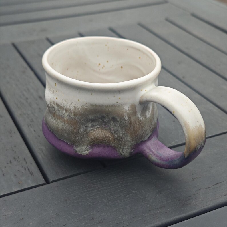 Op de afbeelding: Een handgemaakte keramische mok met een witte binnenkant en een oor. De buitenkant heeft een kleurengradi&euml;nt, van wit naar bruin, grijs en paars aan de basis. De mok staat op een grijs houten oppervlak.