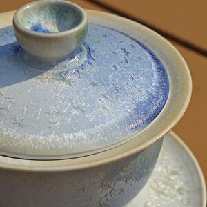 May include: A ceramic tea set with a light blue and white design. The lid and cup have a textured, crystalline pattern. The small lid handle is a contrasting green and blue. The set is on a light brown surface.