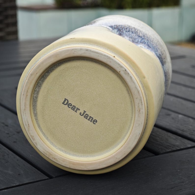 May include: A cylindrical ceramic vessel with a cream-colored base and a blue and white mottled glaze on the upper portion. The bottom of the vessel features a raised circular rim and the text Dear Jane stamped in dark gray lettering.