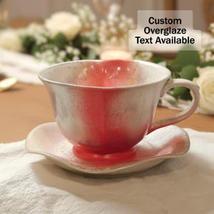 May include: A ceramic teacup and saucer set with a white and red glaze. The cup has a rounded shape with a handle, and the saucer has a scalloped edge. The text "Custom Overglaze Text Available" is visible.