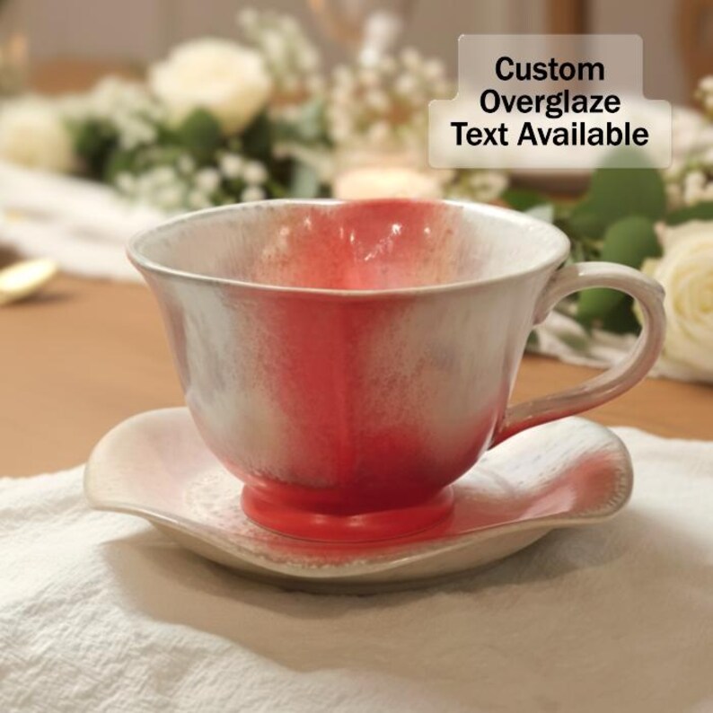 May include: A ceramic teacup and saucer set with a white and red glaze. The cup has a rounded shape with a handle, and the saucer has a scalloped edge. The text "Custom Overglaze Text Available" is visible.
