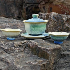May include: A ceramic tea set with a light blue, green, and yellow glaze. The set includes a lidded tea bowl, a saucer, and two small teacups. The pieces are arranged on a textured rock surface.