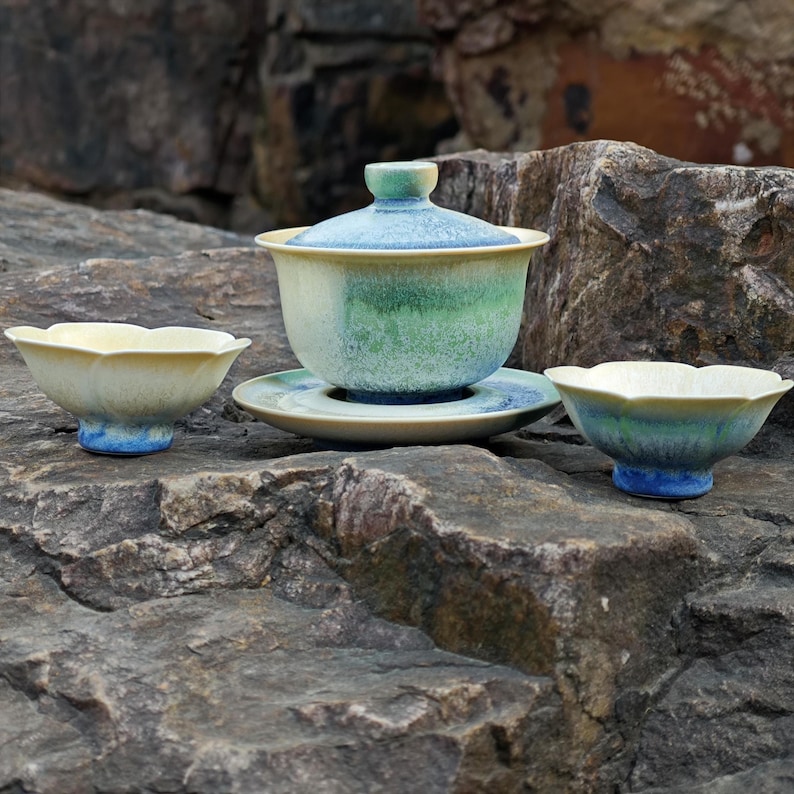 May include: A ceramic tea set with a light blue, green, and yellow glaze. The set includes a lidded tea bowl, a saucer, and two small teacups. The pieces are arranged on a textured rock surface.