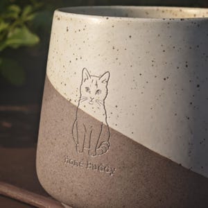 May include: A two-toned ceramic mug featuring a speckled cream upper section and a solid dark grey lower section. An engraved line drawing of a seated cat is centred on the front, positioned above the embossed text Home Buddy.