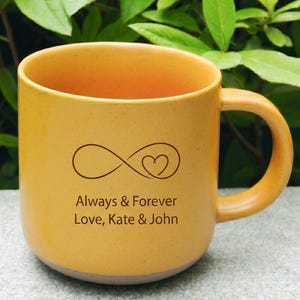 Puede incluir: Taza de cerámica de color amarillo mostaza con asa curva. La taza presenta un símbolo de infinito marrón con un corazón y el texto "Always & Forever Love, Kate & John."