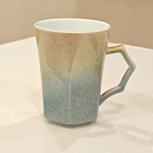 May include: A ceramic mug with a unique geometric handle. The mug features a gradient of colors, transitioning from beige to blue. The interior is white. The mug is designed with a modern aesthetic.
