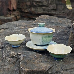 May include: A ceramic tea set with a floral design. The set includes a lidded tea bowl, a saucer, and two small bowls. The colors are primarily cream, blue, and green, with a speckled glaze. The set is displayed on a gray stone surface.