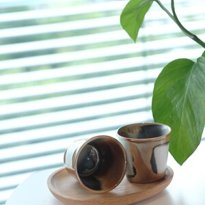 以下が含まれることがあります： 木製のトレイに、茶色、黒、白の抽象的なデザインのセラミックカップが2つあります。カップの内側は濃く、縁は薄茶色です。背景には大きな葉を持つ緑の植物があります。