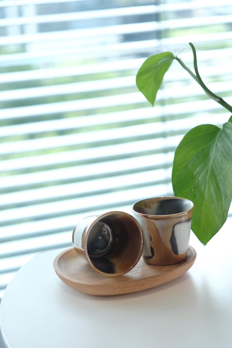 以下が含まれることがあります： 木製のトレイに、茶色、黒、白の抽象的なデザインのセラミックカップが2つあります。カップの内側は濃く、縁は薄茶色です。背景には大きな葉を持つ緑の植物があります。