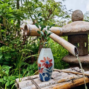May include: A ceramic vase with a floral design, featuring red and blue flowers, holds a green leafy stem. The vase sits on a bamboo structure, with a stone lantern and flowing water in the background. The scene evokes a sense of tranquility.
