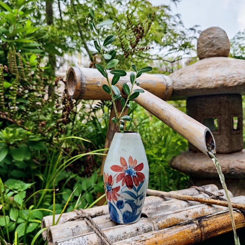 May include: A ceramic vase with a floral design, featuring red and blue flowers, holds a green leafy stem. The vase sits on a bamboo structure, with a stone lantern and flowing water in the background. The scene evokes a sense of tranquility.