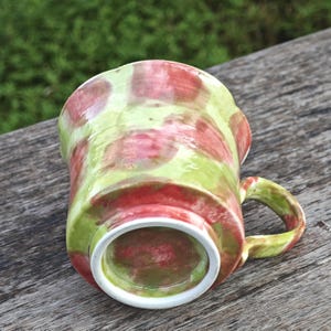 May include: A handmade ceramic mug with a unique design. The mug features a green and red spotted pattern, with a white interior and handle. The mug is set against a natural wood surface, showcasing its handcrafted quality.