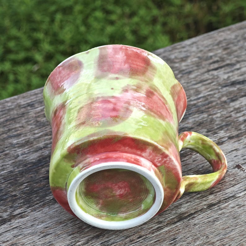 May include: A handmade ceramic mug with a unique design. The mug features a green and red spotted pattern, with a white interior and handle. The mug is set against a natural wood surface, showcasing its handcrafted quality.