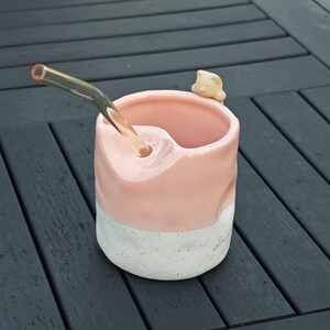 May include: A handmade ceramic mug with a pink upper section and a speckled white base. A clear glass straw extends from the mug, and a small, beige cat figurine rests on the rim. The mug is designed for beverages.
