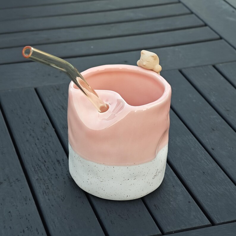 May include: A handmade ceramic mug with a pink upper section and a speckled white base. A clear glass straw extends from the mug, and a small, beige cat figurine rests on the rim. The mug is designed for beverages.