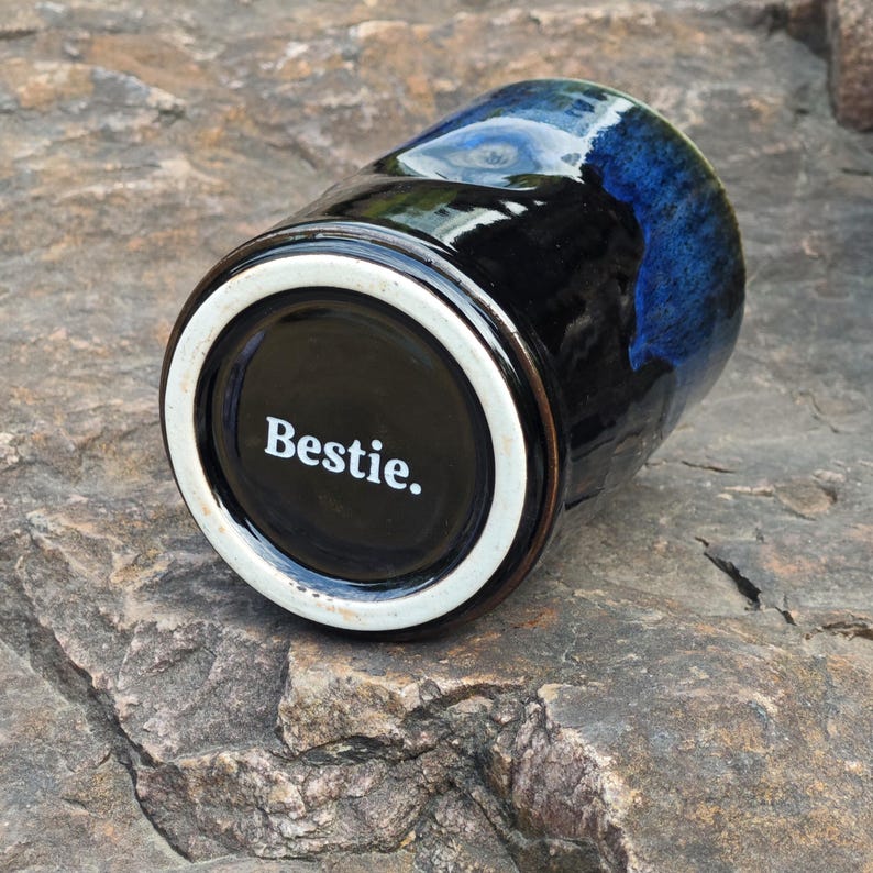 May include: A ceramic mug featuring a glossy black and deep blue reactive glaze finish. The bottom of the mug is white with the word Bestie. printed in a clean white font. The mug is positioned at an angle, showcasing the smooth, reflective surface.