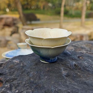 May include: Two ceramic tea bowls, one stacked on the other, with a flower petal design. The top bowl is a creamy beige, while the bottom bowl has a gradient of green and blue. The bowls are on a dark gray rock.
