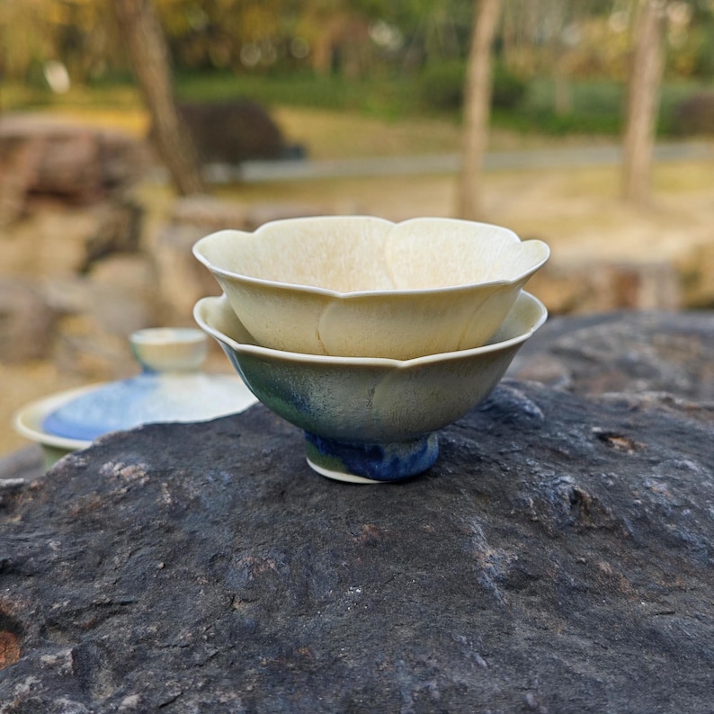 May include: Two ceramic tea bowls, one stacked on the other, with a flower petal design. The top bowl is a creamy beige, while the bottom bowl has a gradient of green and blue. The bowls are on a dark gray rock.