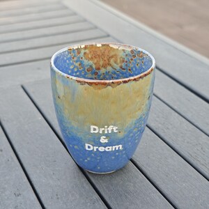 Puede incluir: Un vaso de cer&aacute;mica con un esmalte moteado azul y beige. Las palabras "Drift & Dream" est&aacute;n impresas en blanco en el lateral. El vaso es cil&iacute;ndrico con un borde ligeramente ensanchado.