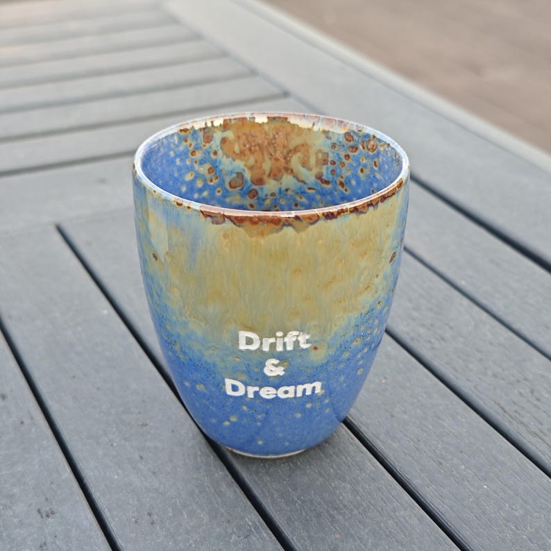 Puede incluir: Un vaso de cer&aacute;mica con un esmalte moteado azul y beige. Las palabras "Drift & Dream" est&aacute;n impresas en blanco en el lateral. El vaso es cil&iacute;ndrico con un borde ligeramente ensanchado.