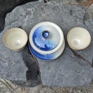 May include: A ceramic tea set consisting of a lidded gaiwan and two matching cups. The pieces feature a gradient glaze transitioning from a deep cobalt blue at the base and lid center to a creamy off-white finish with a smooth, rounded rim.