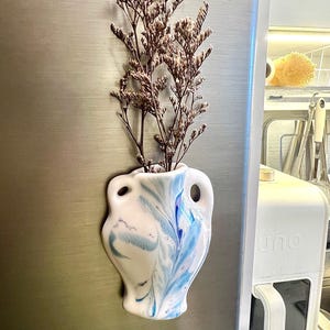 Op de afbeelding: Een witte keramische vaas met blauwe marmeren accenten, gevuld met gedroogde bruine bloemen. De vaas heeft een unieke vorm met twee handvatten en is bevestigd aan een metalen oppervlak, waarschijnlijk een koelkast. Een decoratief item voor in huis.