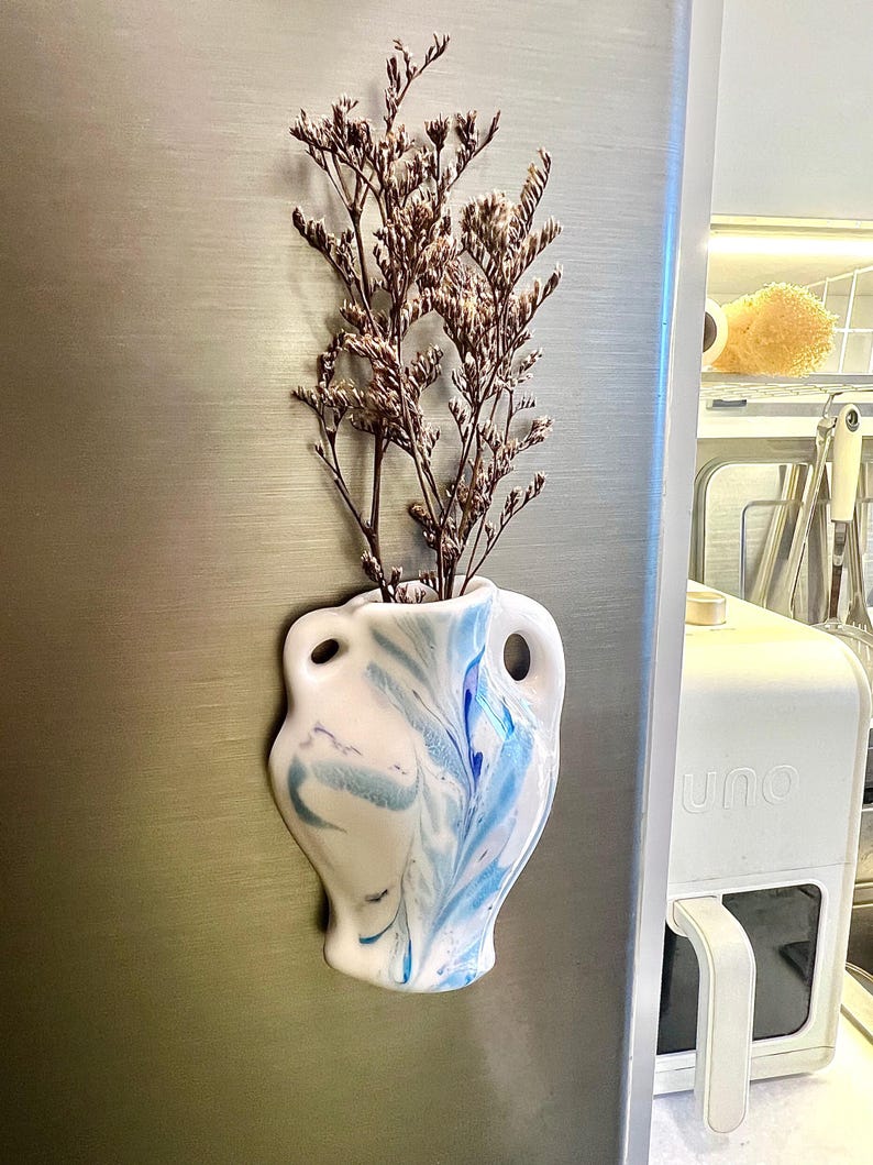 Op de afbeelding: Een witte keramische vaas met blauwe marmeren accenten, gevuld met gedroogde bruine bloemen. De vaas heeft een unieke vorm met twee handvatten en is bevestigd aan een metalen oppervlak, waarschijnlijk een koelkast. Een decoratief item voor in huis.