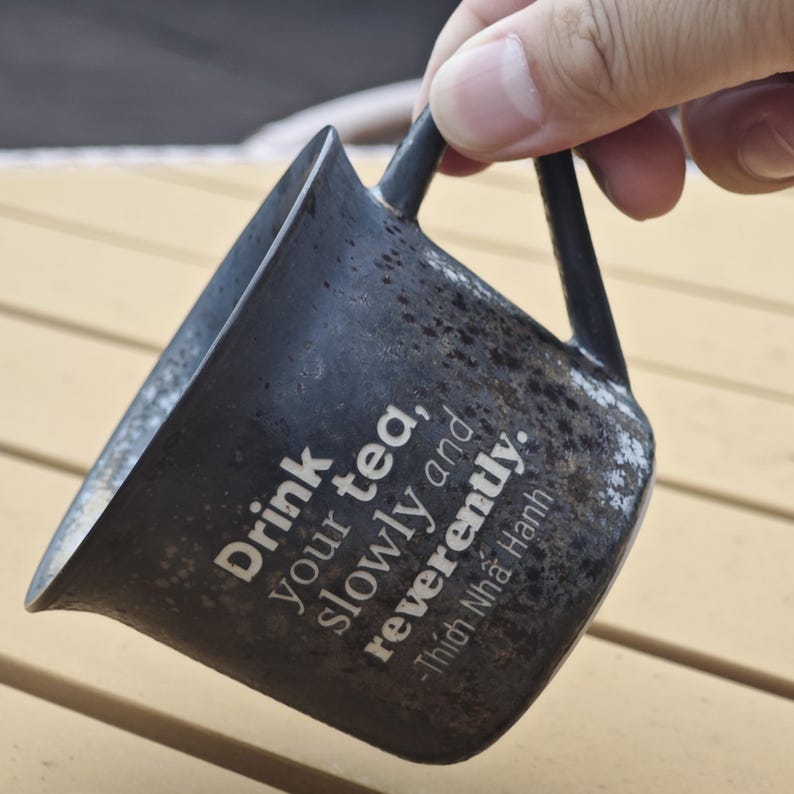 May include: A black ceramic mug with a speckled finish, held by a hand. The mug features the text "Drink your tea, slowly and reverently." and a quote by Thich Nhat Hanh. The mug has a unique, angular design.