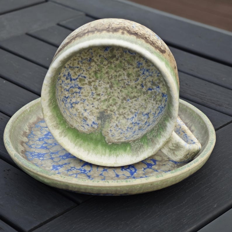 May include: A ceramic teacup and saucer set. The cup is upside down, revealing a speckled interior with blue and green hues. The saucer mirrors the cup's design, with a green rim and a speckled pattern.