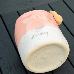 May include: A ceramic mug with a pink upper half and a speckled white lower half. The mug has a tan handle and the word "Lowkey" is written on the side. The mug is sitting on a dark wooden surface.
